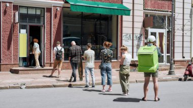 Bekleyen, kuyrukta bekleyen, siparişlerini almak için sosyal mesafeye saygı duyan, Coronavirus tecridi sırasında toplanma noktasından alışveriş yapan insanların tam boy görüntüsü.