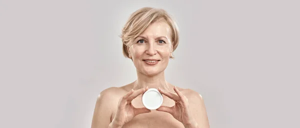 Portrait of beautiful middle aged woman looking at camera, holding moisturizing facial cream while posing isolated over grey background — Stock Photo, Image
