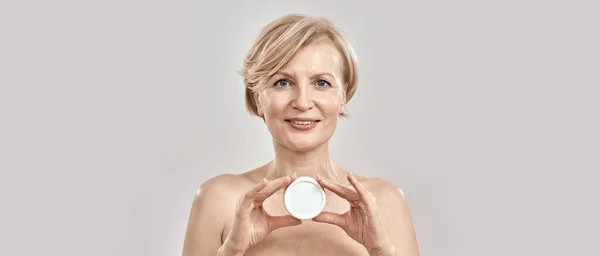 Retrato de bela mulher de meia-idade olhando para a câmera, segurando creme facial hidratante enquanto posando isolado sobre fundo cinza — Fotografia de Stock