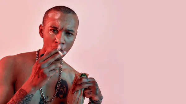Topless, sötét bőrű férfi tetoválással, aki cigarettát szív, cigarettát tart egy üveggel papírzacskóban, és a kamerába néz. — Stock Fotó