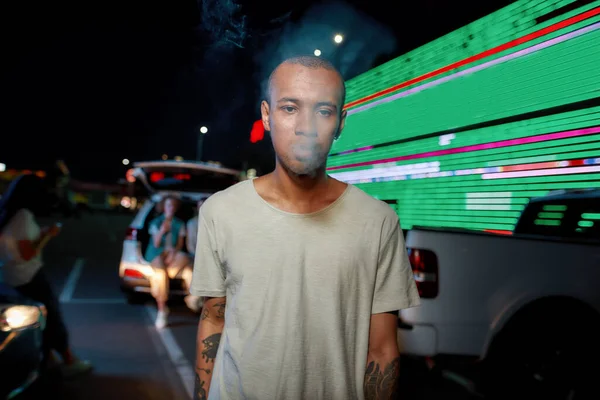 Um close-up de um jovem de pele escura casualmente vestido tatuado homem olhando para uma câmera enquanto estava no meio de uma fumaça ter amigos e uma tela de LED em um fundo — Fotografia de Stock