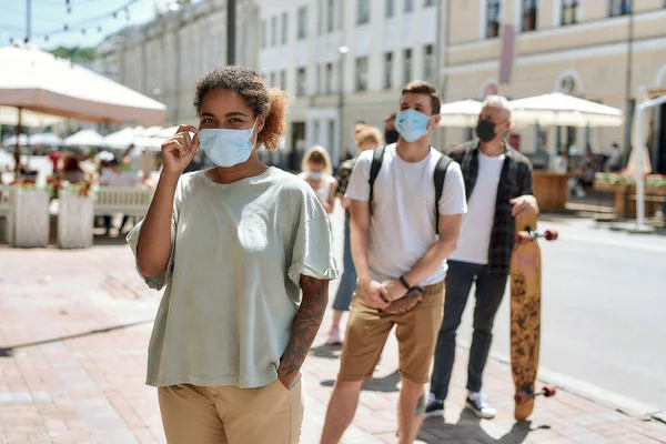 Молодая африканская американка в маске, стоящая в очереди с другими людьми, соблюдая социальное дистанцирование при входе в ресторан на вынос во время блокировки коронавируса — стоковое фото