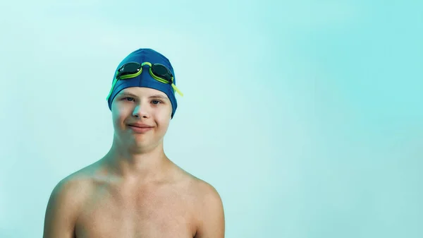 Niño discapacitado con síndrome de Down en gorra de natación con gafas sonriendo a la cámara mientras posa aislado sobre fondo turquesa —  Fotos de Stock