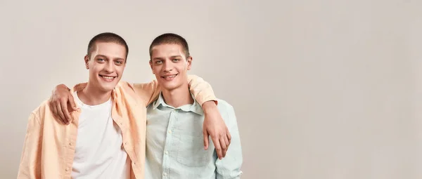 Два счастливых молодых брата-близнеца обнимаются и улыбаются в камеру, позируя вместе, изолированные на бежевом фоне — стоковое фото