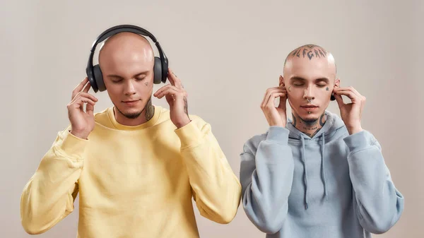 Dois jovens irmãos gêmeos em desgaste casual com tatuagens e piercings ouvindo música em fones de ouvido e fones de ouvido sem fio, de pé isolados em fundo bege — Fotografia de Stock