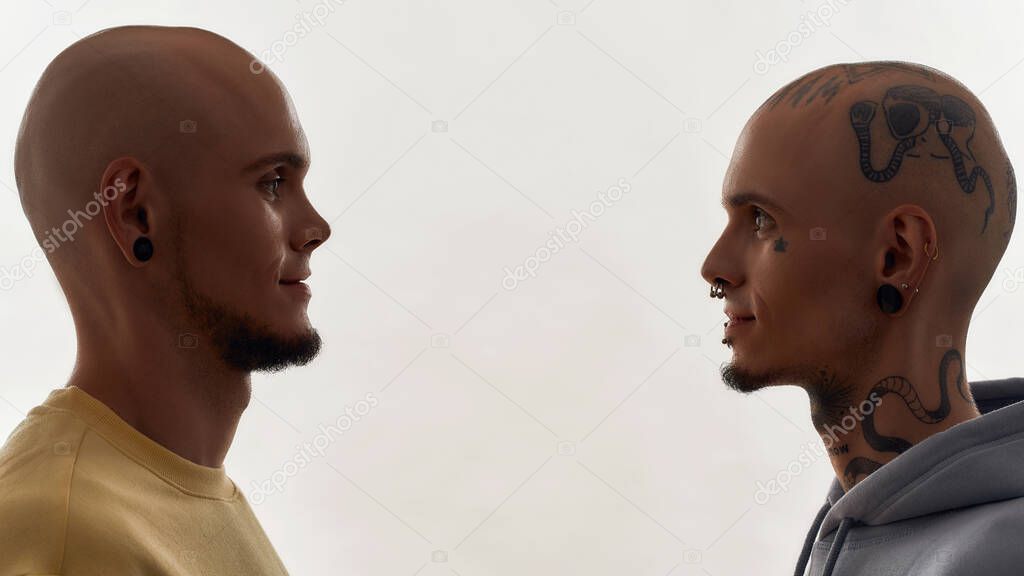 Two young caucasian twin brothers with tattoos and piercings looking at each other while standing face to face isolated over white background