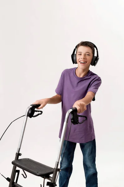 Ευτυχισμένο έφηβο ανάπηρο αγόρι με εγκεφαλική παράλυση στα ακουστικά χαμογελώντας στην κάμερα, κάνοντας βήματα με τον περιπατητή του απομονωμένο σε λευκό φόντο — Φωτογραφία Αρχείου