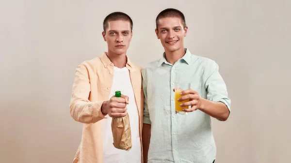 Dois jovens irmãos gêmeos caucasianos em uso casual segurando garrafa de cerveja dentro de saco de papel e vidro de laranja fresco enquanto estava isolado sobre fundo bege — Fotografia de Stock