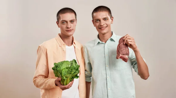 Nutrição vegetariana. Dois jovens irmãos gêmeos caucasianos em uso casual segurando salada verde fresca e pedaço de carne enquanto estavam isolados sobre fundo bege — Fotografia de Stock