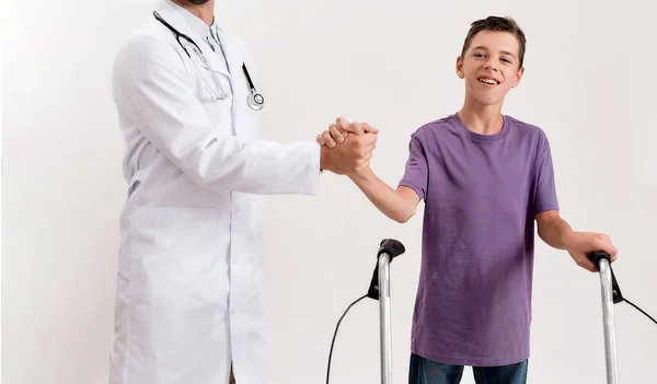 Oříznutý snímek mužského lékaře třesoucí se ruce s dospívajícím postiženým chlapcem s mozkovou obrnou, přičemž kroky pomocí jeho chodítko izolované přes bílé pozadí — Stock fotografie