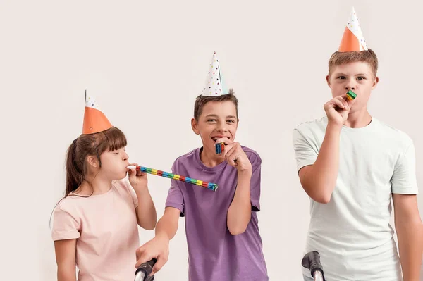 Trójka niepełnosprawnych dzieci z zespołem Downa i porażeniem mózgowym noszących czapki urodzinowe i gwizdki podczas wspólnego świętowania urodzin na białym tle — Zdjęcie stockowe