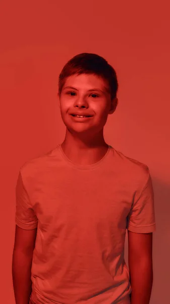 Retrato de adolescente alegre com deficiência menino com síndrome de Down sorrindo para a câmera enquanto posando isolado sobre fundo de luz vermelha — Fotografia de Stock