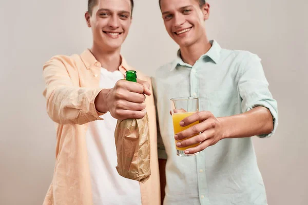 Два молодых белых брата-близнеца в повседневной одежде держат бутылку пива внутри бумажного пакета и стакан апельсина свежим, стоя изолированно на бежевом фоне, сосредоточиться на напитках — стоковое фото