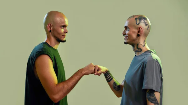Πορτρέτο των νέων δίδυμων αδελφών με τατουάζ και σκουλαρίκια κοιτάζοντας ο ένας τον άλλο, δίνοντας γροθιά χτύπημα, στέκεται πρόσωπο με πρόσωπο απομονώνονται σε ανοιχτό πράσινο φόντο — Φωτογραφία Αρχείου
