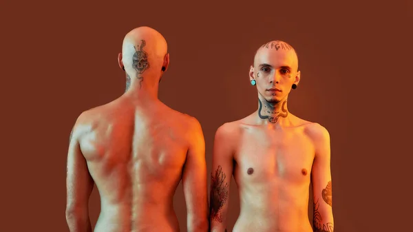 Visão frontal e traseira de jovens meio nus irmãos gêmeos com tatuagens e piercings posando juntos, em pé isolado sobre fundo laranja — Fotografia de Stock