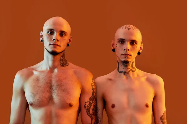 Retrato de jovens meio nus irmãos gêmeos com tatuagens e piercings olhando para a câmera, posando juntos, de pé isolados sobre fundo laranja — Fotografia de Stock