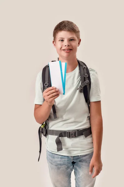 Niño discapacitado feliz con síndrome de Down usando mochila sonriendo mientras sostiene boletos aéreos, de pie aislado sobre fondo blanco —  Fotos de Stock