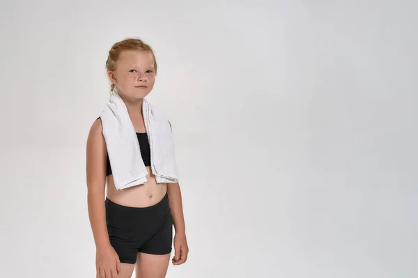 Όμορφη μικρή κοκκινομάλλα σπορ κορίτσι στο αθλητικό ντύσιμο κοιτάζοντας κάμερα, στέκεται με λευκή πετσέτα γύρω από το λαιμό της, ενώ θέτουν απομονώνονται σε λευκό φόντο — Φωτογραφία Αρχείου
