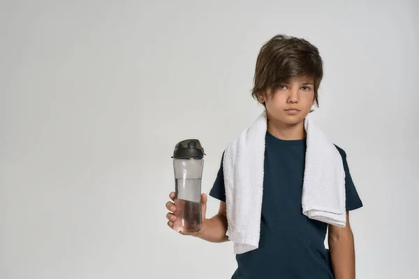 Μικρό σπορ παιδί με αθλητικά ρούχα που κοιτάζει την κάμερα, κρατώντας ένα μπουκάλι νερό ενώ στέκεται με λευκή πετσέτα γύρω από το λαιμό του απομονωμένο σε λευκό φόντο — Φωτογραφία Αρχείου