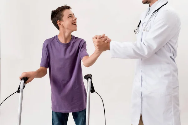 Cortado tiro de médico masculino apertando as mãos com menino deficiente alegre com paralisia cerebral, tomando medidas usando seu andador isolado sobre fundo branco — Fotografia de Stock