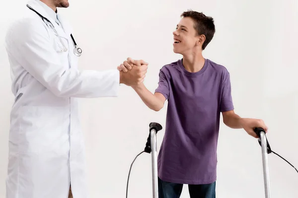 Cortado tiro de médico masculino apertando as mãos com menino deficiente alegre com paralisia cerebral, tomando medidas usando seu andador isolado sobre fundo branco — Fotografia de Stock