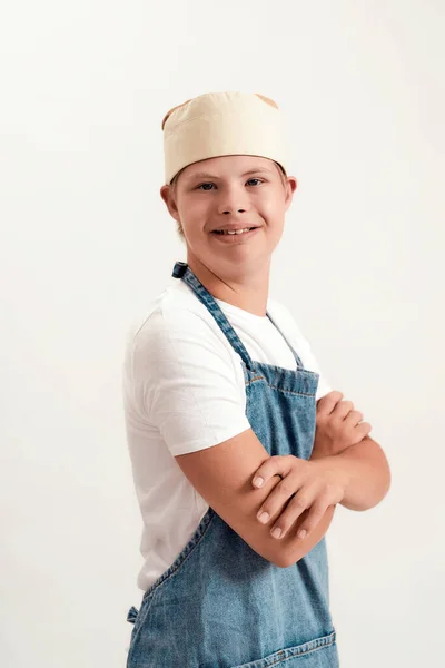 Retrato de menino com deficiência com síndrome de Down vestido como um cozinheiro de avental e chapéu sorrindo para a câmera enquanto de pé com os braços cruzados isolado sobre fundo branco — Fotografia de Stock