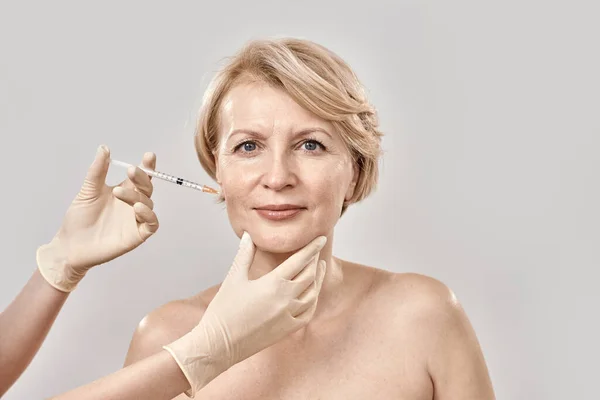 Retrato de bela mulher de meia-idade nua olhando para a câmera enquanto ela recebe injeção em seu rosto, posando isolado contra fundo cinza — Fotografia de Stock