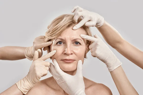 Beauty portrait of middle aged woman going under cosmetic treatment. Beautician hands in gloves checking female face skin isolated against grey background — Zdjęcie stockowe