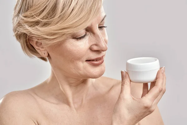 Close up portrait of beautiful middle aged woman holding, looking at moisturizing cream jar while posing isolated over grey background — Stock Photo, Image