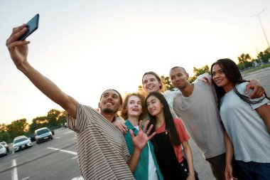 Bir grup genç ortak giyinmiş arkadaş birlikte iyi vakit geçiriyorlar. Birbirlerine sarılıyorlar ve arka planda arabaları olan bir otoparkta grup selfie çekiyorlar.
