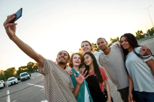 Un grupo de seis jóvenes amigos vestidos comúnmente pasar un buen rato juntos abrazándose y haciendo un selfie grupo fuera en un sitio de estacionamiento con coches en un fondo —  Fotos de Stock