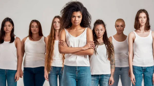 Retrato de jovem afro-americana de camisa branca e jeans jeans posando com braços cruzados. Grupo de mulheres diversas de pé isolado sobre fundo cinza — Fotografia de Stock