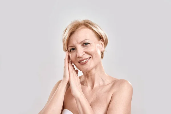 Portrait of beautiful middle aged woman touching her skin, smiling at camera while posing isolated against grey background. Beauty concept — Stock Photo, Image