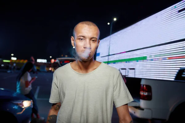 A young dark-skinned well-dressed tattooed man looking into a camera while breathing out smoke having friends and a led screen on a background