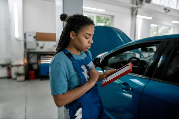 Excellentie van voertuigen. Jonge Afrikaanse Amerikaanse vrouw, professionele vrouwelijke monteur met behulp van tablet pc, terwijl staan in de buurt van blauwe auto met open motorkap bij auto reparatiewerkplaats — Stockfoto