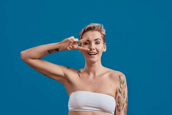Ritratto di una giovane attraente donna mezza nuda tatuata con pelle perfetta dall'aspetto felice, che tiene la mano sugli occhi e mostra vittoria o segno di pace isolato su sfondo blu — Foto Stock