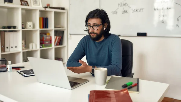 Jeune professeur homme barbu assis à son lieu de travail et enseignant l'informatique en ligne. Regarder webcam et parler avec le public — Photo