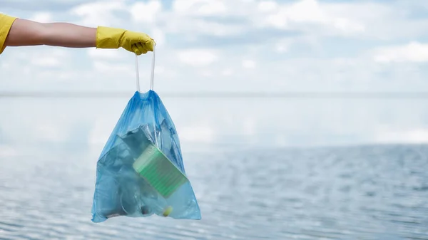 Limpiando playa. Voluntarios mano en guante de goma amarilla sosteniendo bolsa de basura con residuos de plástico y otra basura contra el mar y el cielo — Foto de Stock