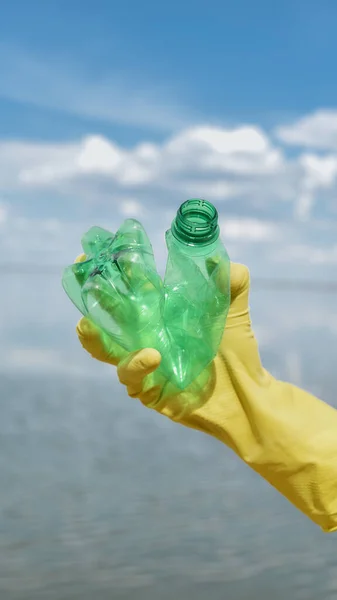 Reciclaje. Tiro vertical de un voluntario con guante de basura amarillo sosteniendo botella de plástico usado contra el agua, el río o el lago — Foto de Stock