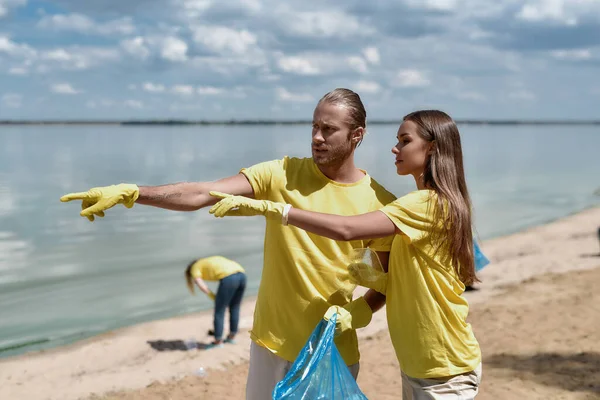 Чистая вода. Два молодых эко-активиста, мужчина и женщина, держат мешок для мусора и обсуждают что-то во время уборки пляжной зоны из пластика с командой добровольцев — стоковое фото