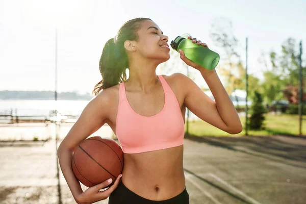 Happy young female basketball player smiling aside while drinking water from bottle, holding basketball, standing outdoors on a sunny day