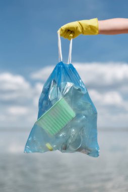 Dikey eko aktivistin sarı lastik eldiven giyip plastik atık dolu çöp torbası tutarken sahili ve gökyüzünü temizlerken görüntüsü.