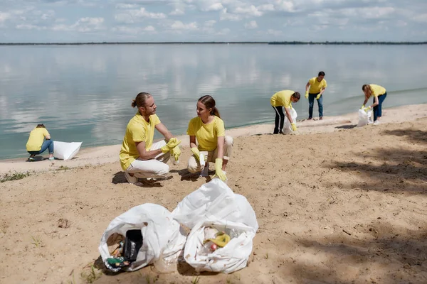 Salva nuestro planeta. Grupo de jóvenes eco activistas o voluntarios en uniforme limpiando juntos la playa de la recolección de residuos plásticos y basura, dos bolsas de basura en la arena — Foto de Stock