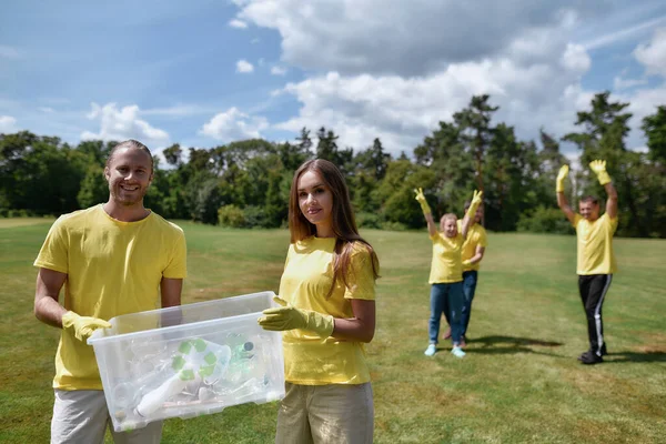 Двое молодых эко-активистов в форме держат мусорный бак с пластиковыми бутылками во время уборки парка вместе с группой волонтеров. — стоковое фото