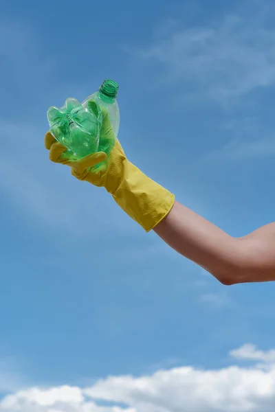 Recykling. Pionowe ujęcie ochotnika w żółtej rękawiczce na śmieci z plastikową butelką na tle błękitnego nieba — Zdjęcie stockowe