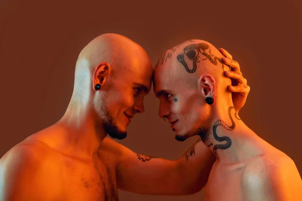 Porträt junger Zwillingsbrüder mit Tätowierungen und Piercings, die Kopf an Kopf stehen und einander isoliert auf orangefarbenem Hintergrund anlächeln — Stockfoto