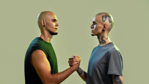 Ritratto di giovani fratelli gemelli con tatuaggi e piercing che si guardano, si tengono per mano, in piedi faccia a faccia isolati su sfondo verde chiaro — Foto Stock