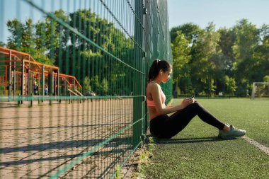 Genç sporcu kadın kulaklık takıyor açık havada oturuyor, müzik dinliyor, akıllı telefondan çalma listesine bakıyor ve gülümsüyor.