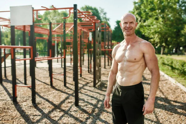 Pria dewasa berotot, binaragawan dengan tubuh telanjang tersenyum di depan kamera, berpose di luar ruangan saat latihan di lapangan olahraga jalanan — Stok Foto