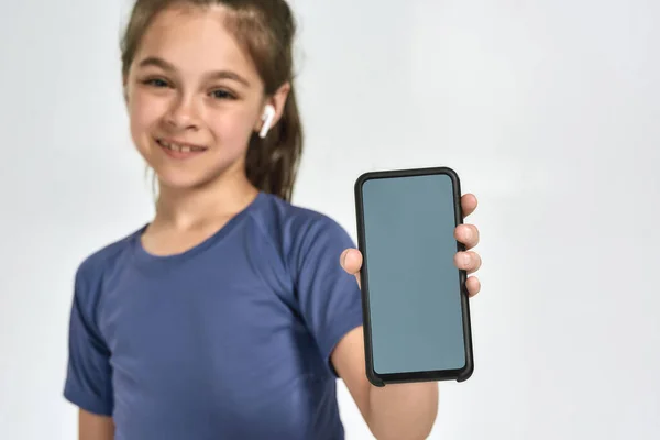 Pequena criança menina esportiva usando fones de ouvido, sorrindo para a câmera e mostrando smartphone com tela em branco, de pé isolado sobre fundo branco — Fotografia de Stock